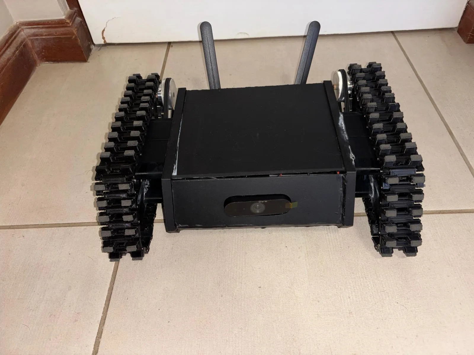 Photograph of the finished robot on a tiled floor: a black box body between two tank tracks, a webcam in the front panel and two antennas at the back